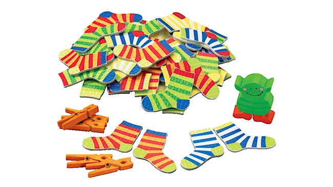 Das Bild zeigt das Kinderspiel "Socken zocken". Es besteht aus einer Vielzahl bunter, gestreifter Sockenpaare, einigen orangefarbenen Wäscheklammern und einer grünen Spielfigur in Form eines Monsters. Die Socken sind in verschiedenen Farben und Mustern gestaltet und liegen verstreut auf einer Fläche.