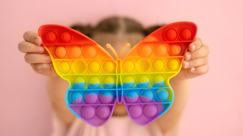 Ein Mädchen hält ein leuchtend buntes Pop It-Spielzeug in Form eines Schmetterlings vor ihr Gesicht vor einem rosa Hintergrund. 
