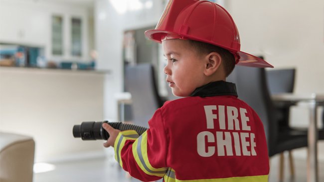 Gezeigt wird ein kleiner Junge in einem Feuerwehrmann-Kostüm, der einen Schlauch zum Löschen in der Hand hält. Er steht in einer modern eingerichteten Wohnung. Im Hintergrund sind ein Küchentresen und ein Esstisch mit Stühlen zu sehen. Die Ecke eines Sofas ragt unten in der linken Ecke ins Bild.