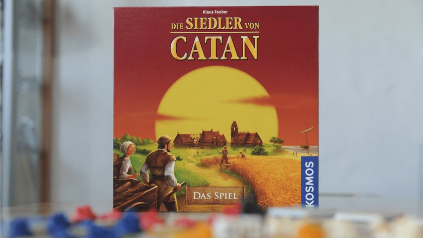 Gezeigt wird die Box des Bettspiels Catan, wie sie 2013 aussah. Die Box ist rostrot, im Hintergrund ist ein großer Sonnenuntergang und ein Dorf zu sehen, auf das ein Weg zuführt. Links sind grüne Weiden zu sehen, rechts ein Kornfeld, in dem ein Bauer arbeitet. Im Vordergrund ist ein Bauernpaar zu sehen, das über die Feldweg geht. Vor der Box liegen Spielsteine, die nur verschwommen zu sehen sind.