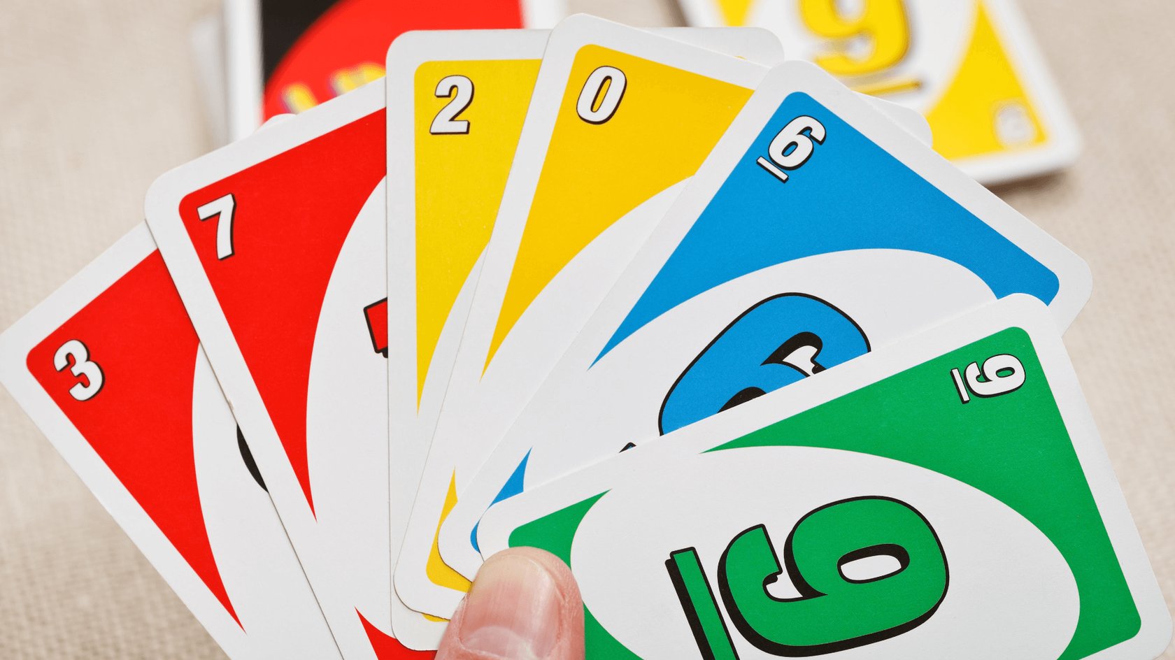 Eine Hand hält mehrere UNO-Karten in den Farben Rot, Gelb, Blau und Grün. Die sichtbaren Zahlen auf den Karten sind 3, 7, 2, 0, 6 und 9. Im Hintergrund sind weitere UNO-Karten zu sehen.