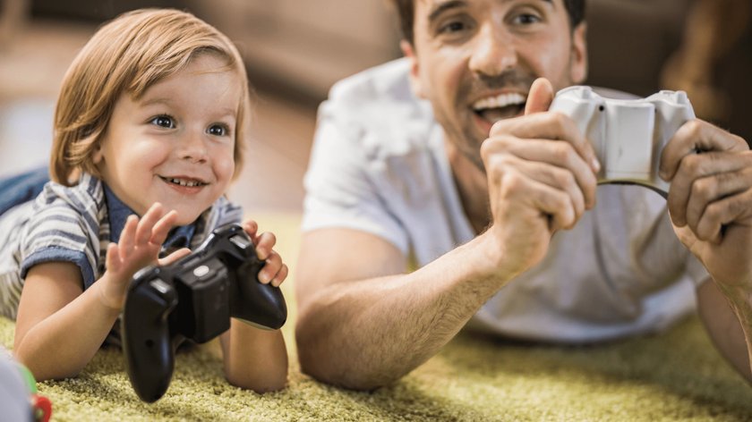 Gezeigt werden ein Vater und sein kleiner Sohn, die bäuchlings auf einem grünen Teppich liegen und je einen PlayStation-Controller in der Hand haben. Beide lachen.