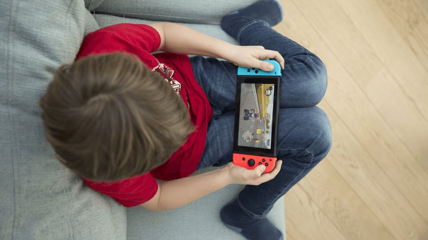 Gezeigt wird ein Junge von ca. 8 Jahren, der auf einem Sofa sitzt und eine Nintendo Switch Konsole in der Hand hält, auf der er das Rennspiel Mario Kart spielt.