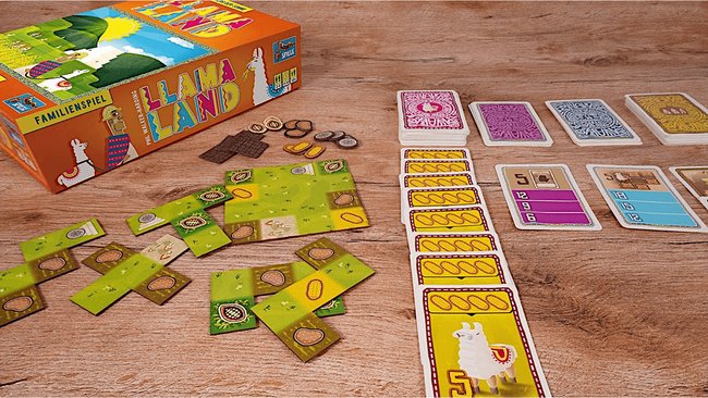 Gezeigt wird die Box und das Spielmaterial des Brettspiels Lamaland. Verschiedene Karten mit Lamas oder Punkten liegen auf einem Tisch. Daneben befinden sich bunte Spielplättchen mit verschiedenen Pflanzen.