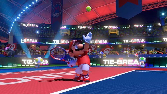 Gezeigt wird ein Screenshot aus dem Switch-Spiel Mario Tennis Aces, in dem Mario auf dem Tennisplatz in einer Halle steht und einen Aufschlag mit dem Tennisball macht.