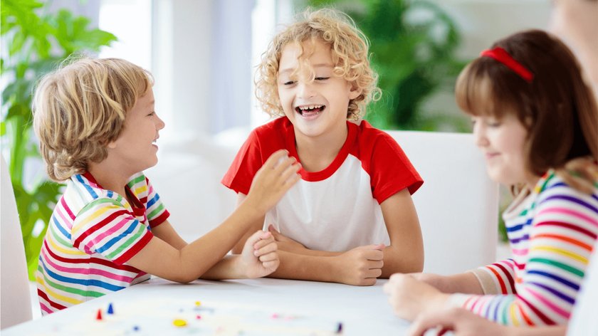 Gezeigt werden drei Kinder, zwei Jungen und ein Mädchen, im Alter von ca. drei bis sieben Jahre, die um einen Tisch sitzen, auf dem ein Gesellschaftsspiel liegt. Zwei der Kinder tragen bunt geringelte T-Shirts. Schemenhaft ist eine ältere Frau am rechten Bildrand zu erkennen, die auf das Spiel schaut.