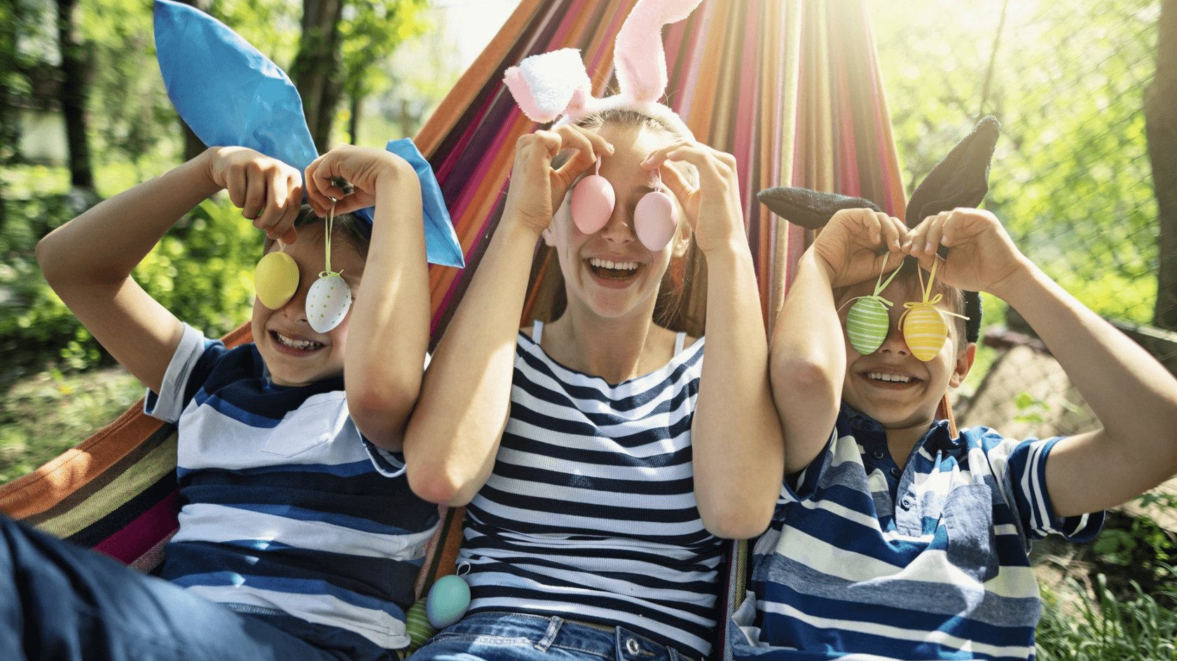 Gezeigt werden drei lachende Kinder in einer Hängematte, die Hasenohren tragen und sich bunte Eier vor die Augen halten. Im Hintergrund sind grüne Bäume und Sonnenschein zu sehen.
