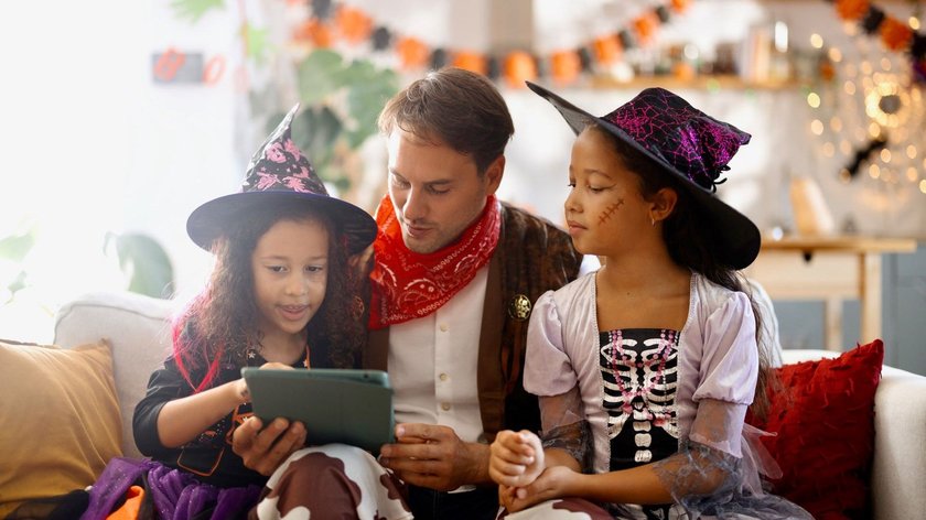 Zwei Mädchen und ein Mann in Halloween-Kostümen sitzen auf einer Couch und schauen auf ein Smartphone. Im Hintergrund sieht man die Dekoration für eine Halloween-Party.