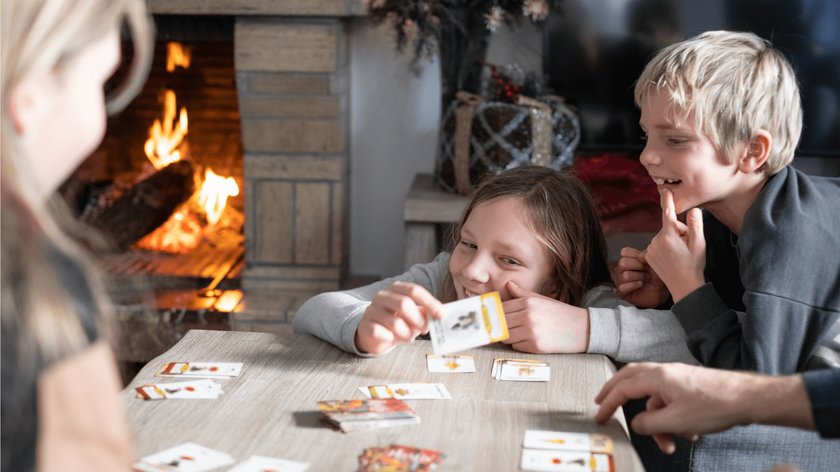 Gezeigt wird eine Familie, die ein Kartenspiel an einem Tisch vor einem Kamin und weihnachtlicher Dekoration spielt.