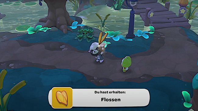 Ein Screenshot aus einem Videospiel zeigt zwei cartoonartige Charaktere auf einem gepflasterten Weg in einer grünen, natürlichen Umgebung mit Pflanzen und Wasser. Ein Schaf-Charakter hält goldene Schwimmflossen. Unten im Bild erscheint eine Textbox mit der Nachricht: "Du hast erhalten: Flossen". Der Hintergrund zeigt einen Baum, Pflanzen und eine kleine Brücke.