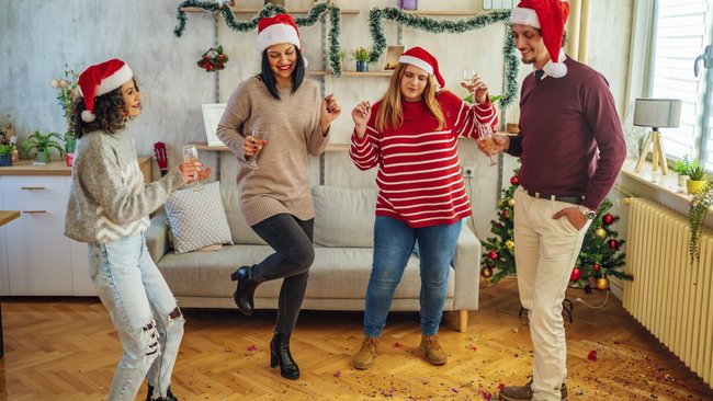 Gezeigt werden vier junge Erwachsene, die mit Weihnachtsmannmützen in einer weihnachtlich dekorierten Wohnung tanzen.