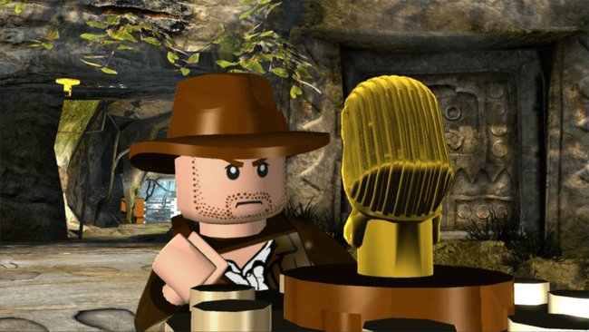 Das Bild zeigt eine Szene aus dem Videospiel LEGO Indiana Jones: Die legendären Abenteuer. Im Vordergrund steht die LEGO-Figur von Indiana Jones mit seinem charakteristischen braunen Hut und Bartschatten, der einen besorgten Gesichtsausdruck zeigt. Neben ihm befindet sich ein goldener Götze oder eine goldene Statue auf einem braunen Sockel, die an die berühmte Eröffnungsszene aus "Jäger des verlorenen Schatzes" erinnert. Im Hintergrund sind steinerne Tempelwände mit Hieroglyphen und ein Eingang zu sehen. Die Szene spielt in einer Höhle oder einem alten Tempel und ist im typischen LEGO-Videospiel-Stil gestaltet.