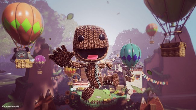 Ein fröhlicher, gestrickter Charakter, bekannt als Sackboy, springt in die Luft und winkt. Im Hintergrund sind mehrere bunte Heißluftballons zu sehen, die über einer malerischen Landschaft mit Häusern und Gärten schweben. Die Szene ist lebendig und spielerisch, mit klaren Details und freundlicher Atmosphäre.