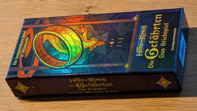 Gezeigt wird die Box des Kartenspiels "Der Herr der Ringe: Die Gefährten – Das Stichspiel" im Querformat. Der Eine Ring aus dem Roman ist auf dem oberen Teil der Box, hier links, groß dargestellt und leuchtet je nach Lichteinfall in bunten Farben, die als Effektfolie über der goldenen Farbe des Rings angebracht sind. Zu sehen sind auch der Spieltitel und keine Informationen wie der Autorname Bryan Bornmueller.