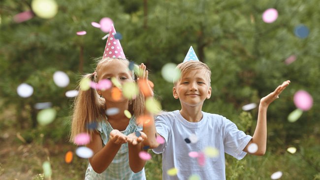 Gezeigt werden zwei Kinder auf einer sommerlichen Wiese, die Konfetti Richtung des Betrachters pusten.