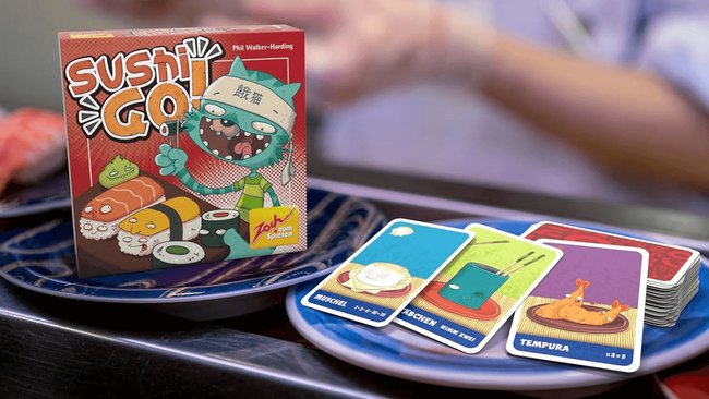 Gezeigt werden die Schachtel und Spielkarten des Kartenspiels Sushi Go auf zwei kleinen Sushi-Tellern.