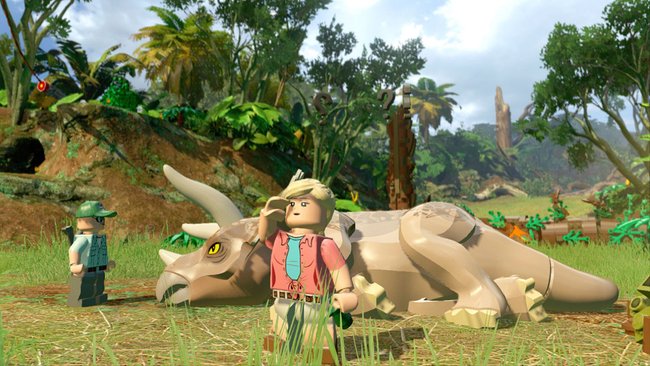 Das Bild zeigt eine Szene aus dem LEGO Jurassic World Videospiel. Im Vordergrund steht eine weibliche LEGO-Figur mit blonden Haaren, die ein rosa Hemd mit türkisfarbener Krawatte trägt. Sie schaut in die Ferne und steht neben einem liegenden LEGO-Triceratops-Dinosaurier in beiger Farbe. Links im Bild ist eine weitere LEGO-Figur mit grünem Hut zu sehen, vermutlich ein Parkwärter oder Wissenschaftler. Die Szene spielt in einer üppigen Dschungelumgebung mit Palmen, Bäumen und grüner Vegetation unter einem leicht bewölkten Himmel. Die Grafik ist im typischen LEGO-Videospiel-Stil gehalten.