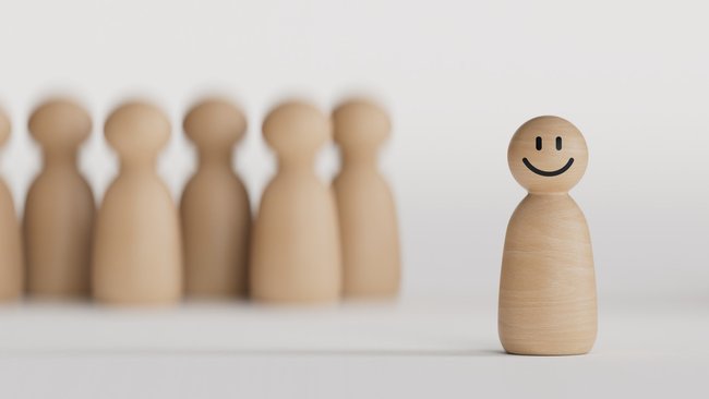 Gezeigt wird eine lachende Spielfigur aus Holz, die sich von den anderen im Hintergrund abhebt.