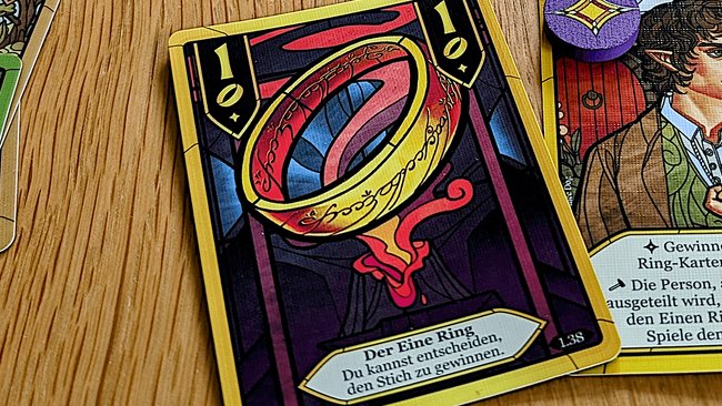 Gezeigt wird die Ring 1-Karte mit dem goldenen Einen Ring mit Elbenschrift aus dem Der Herr der Ringe-Stichspiel, die den Text aufweist: "Der Eine Ring. Du kannst entscheiden, den Stich zu gewinnen."