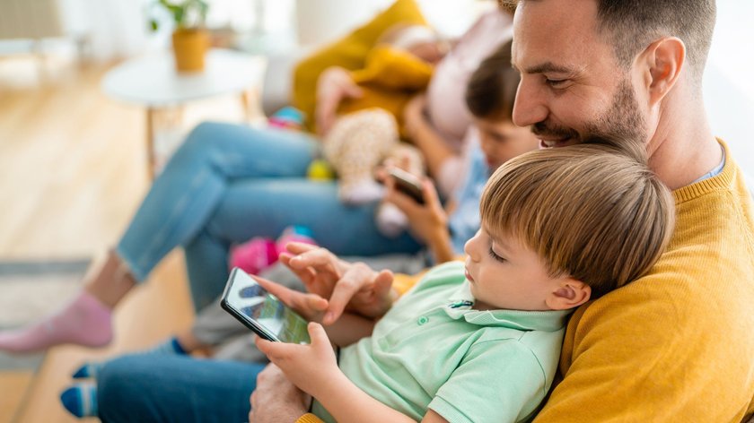 Ein Vater sitzt mit seinem Sohn auf dem Schoß im Wohnzimmer auf der Couch und spielt ein Spiel auf einem Tablet. Im Hintergrund sieht man die restliche Familie, Mutter und Kinder. 