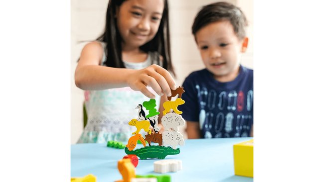 Zwei Kinder spielen mit dem Stapelspiel "Tier auf Tier", bei dem sie bunte Holztiere aufeinander stapeln. Das Spielzeug besteht aus verschiedenen Tierfiguren wie Schafen, Pinguinen und Krokodilen. Die Tiere sind auf einem Tisch angeordnet, und eines der Kinder platziert gerade ein weiteres Tier auf den Stapel.