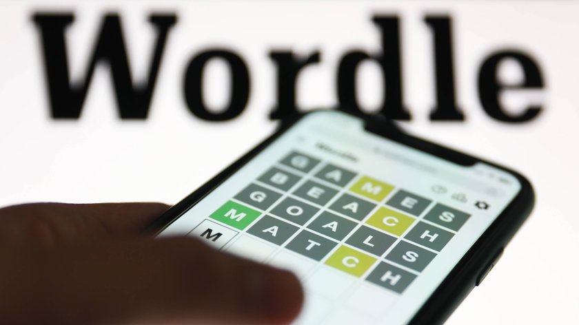 Im Vordergrund sieht man die linke Hand einer Person, die Wordle auf einem Smartphone spielt. Im Hintergrund ist das Wordle-Logo abgebildet.  