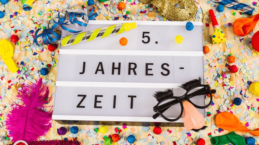 Gezeigt wird eine Tafel mit dem Schriftzug "5.Jahreszeit", die von Konfetti, Luftschlangen, Luftballons ohne Luft, einer Scherzbrille und bunten Federn umgeben ist.