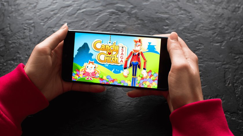 Gezeigt werden zwei Frauenhände, die ein Smartphone halten, auf dem der Startbildschirm der Spiele-App Candy Crush Saga zu sehen ist.