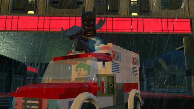 Das Bild zeigt eine Szene aus dem Videospiel LEGO Batman 2: DC Super Heroes. Im Vordergrund ist ein weißer LEGO-Krankenwagen oder Rettungswagen mit roten Akzenten zu sehen, in dem eine LEGO-Figur mit grünen Haaren (vermutlich der Joker) erkennbar ist. Auf dem Dach des Fahrzeugs steht die LEGO-Batman-Figur in seinem charakteristischen schwarzen Kostüm mit gelbem Batman-Logo. Die Szene spielt bei Regen, was durch die sichtbaren Regentropfen dargestellt wird. Im Hintergrund ist ein Gebäude mit roten Leuchtelementen zu erkennen. Die Grafik ist im typischen LEGO-Videospiel-Stil gehalten.