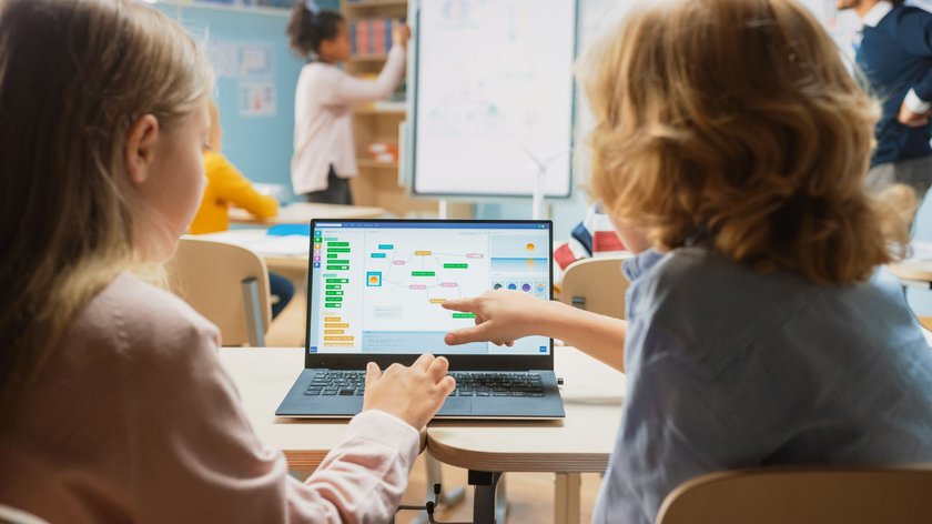 Ein Junge und ein Mädchen schauen in einem Klassenzimmer auf den Bildschirm eines Laptops, der vor ihnen auf dem Tisch steht. Der Junge zeigt auf den Screen, auf dem ein buntes Diagramm zu sehen ist. Im Hintergrund sieht man ein Whitebord und weitere Schülerinnen und Schüler. 