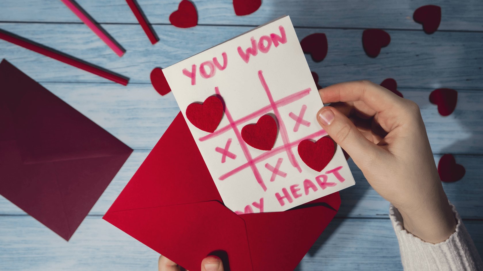 Gezeigt wird eine Karte zum Valentinstag mit einem pinken und roten Tic Tac Toe-Spiel mit Herzen und Kreuzen. Im Hintergrund sind ein roter Briefumschlag, rote und pinke Stifte, rotes Papier und rote Herzen zu sehen.