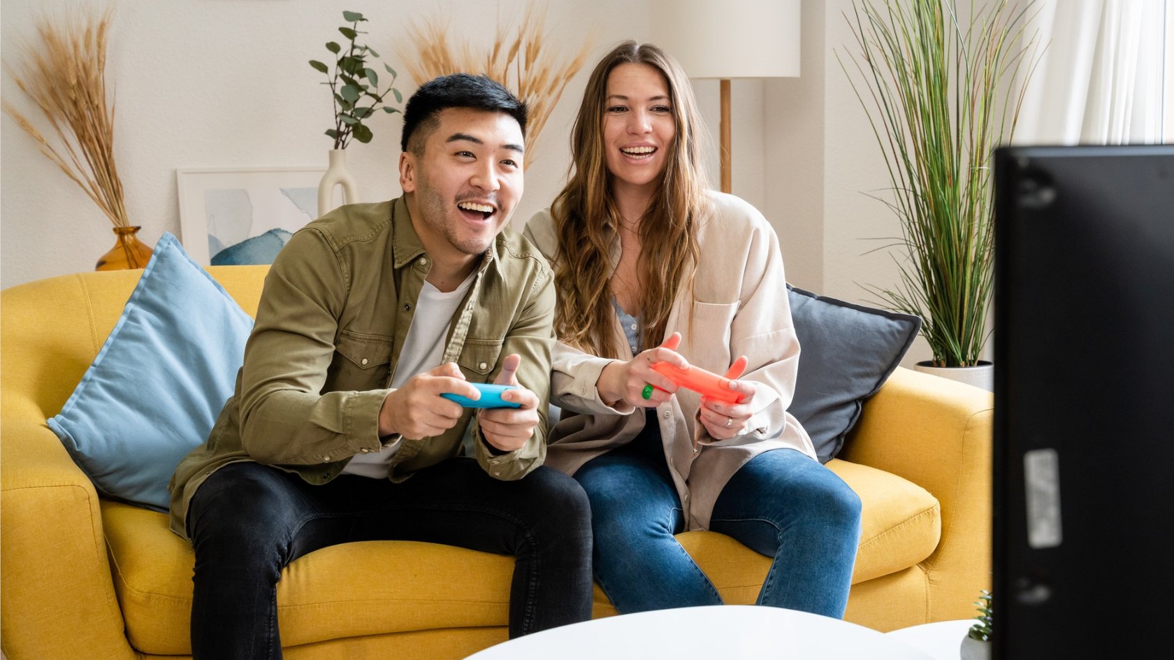 Ein Mann und eine Frau sitzen auf einem gelben Sofa und halten einen blauen und einen roten Spielcontroller, während sie auf einen Fernseher vor ihnen schauen. Im Hintergrund sind eine Zimmerpflanze, ein Kunstwerk und dekorative Zweige in einer Vase zu sehen.