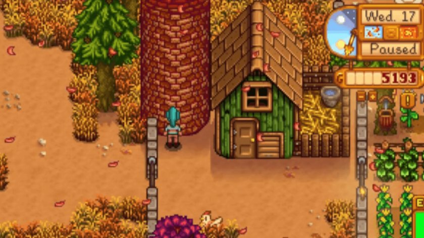 Das Bild zeigt eine Szene aus dem Videospiel Stardew Valley. Im Mittelpunkt steht ein grünes Gebäude mit einem braunen Dach, das an einen Hühnerstall erinnert, daneben ein hohes rotes Silo. Die Umgebung ist herbstlich gestaltet, mit buntem Laub auf dem Boden und Bäumen in warmen Farben. Ein Holz- und Steinfzaun grenzt den Bereich ab. Rechts oben im Bild sind die Spielfortschritt-Anzeigen zu sehen, einschließlich des aktuellen Tages, der Jahreszeit und der Geldanzeige.