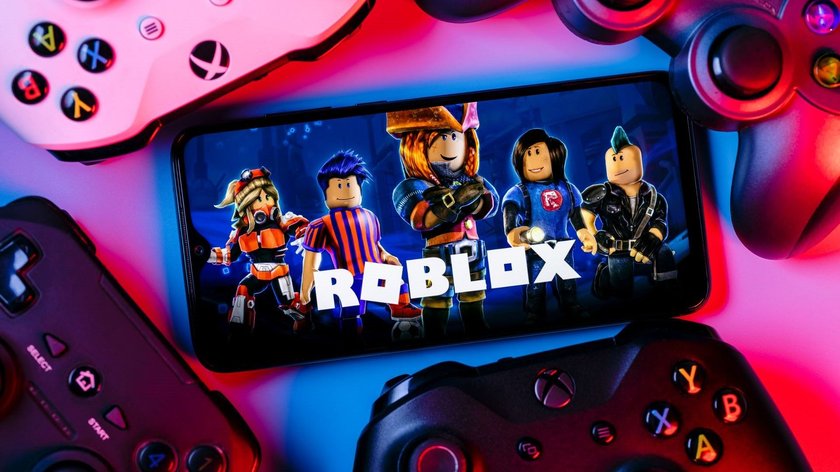 Ein Smartphone liegt in der Mitte zwischen Konsolen und zeigt den Schriftzug Roblox und mehrere Spielfiguren.