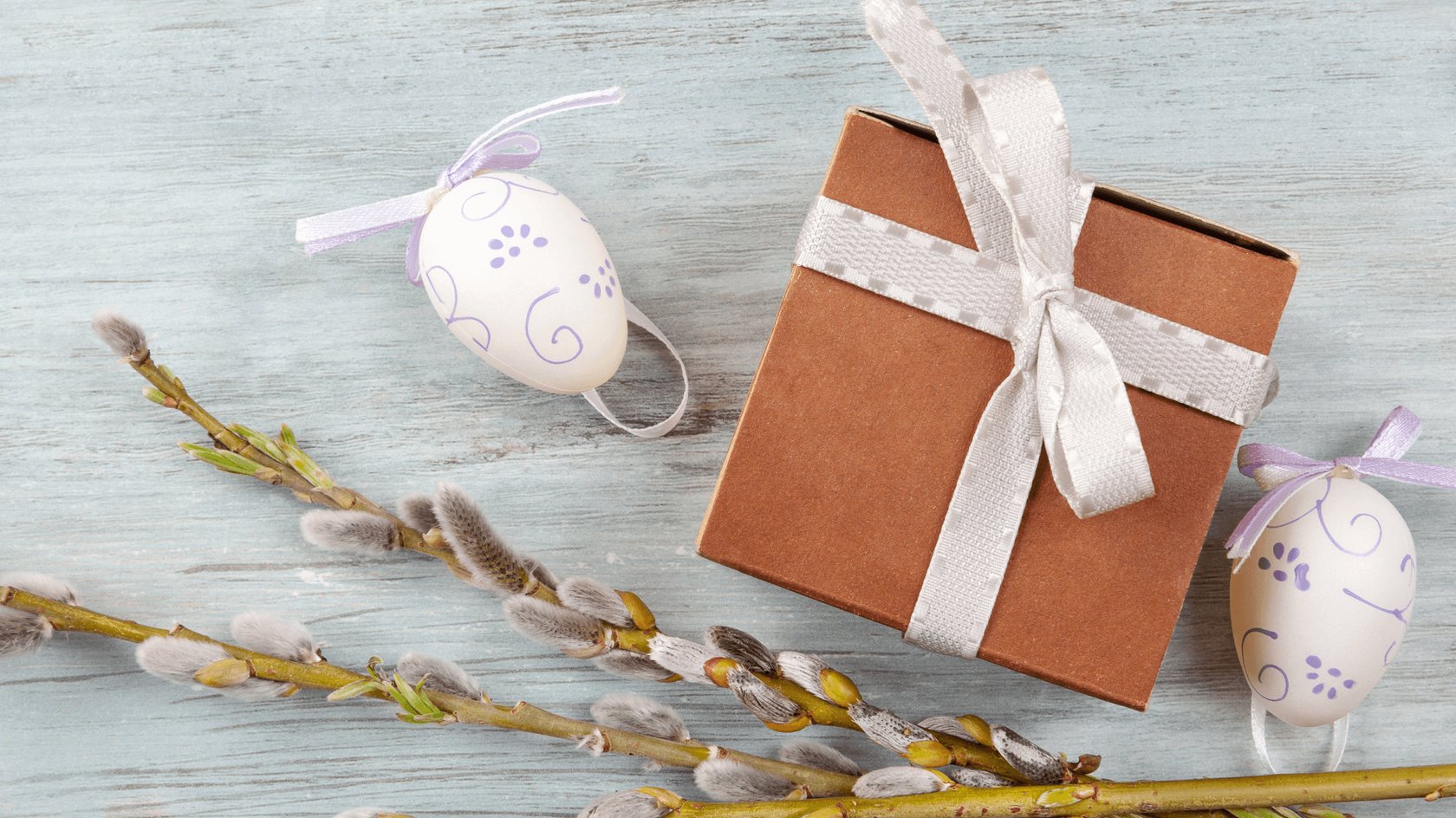 Auf einer hellblauen Holzoberfläche liegen eine braune Geschenkbox mit weißer Schleife, zwei weiße Ostereier mit lila Verzierungen und Schleifen sowie ein Weidenzweig mit flauschigen Kätzchen.